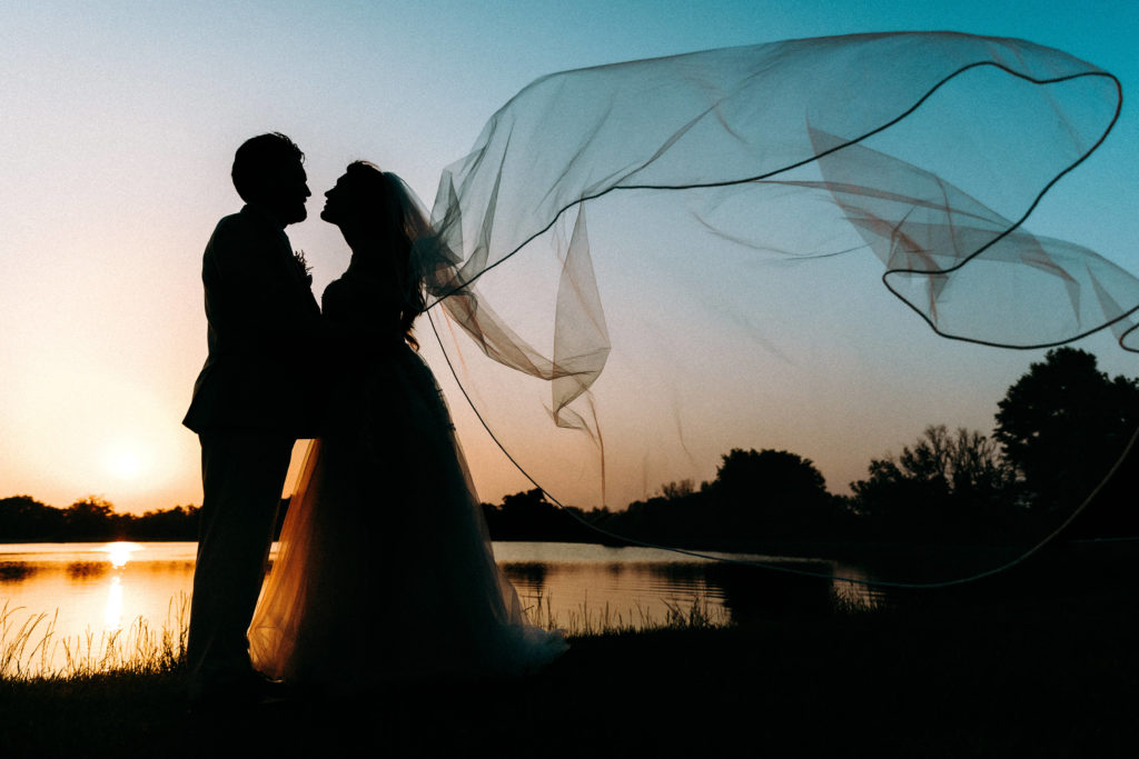Sunset silhouette of a couple with the veil caught in the wind, next to a pond and forest.